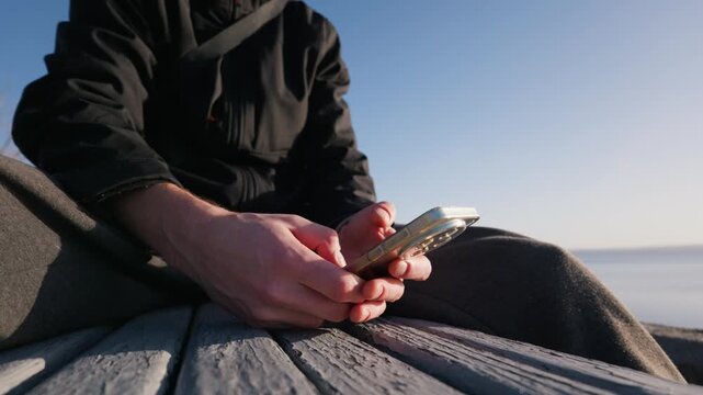 Male hands hold and scroll on a modern smartphone while sitting outdoors. The person is resting cross-legged on a weathered wooden pier by the water under a bright clear blue sky.
