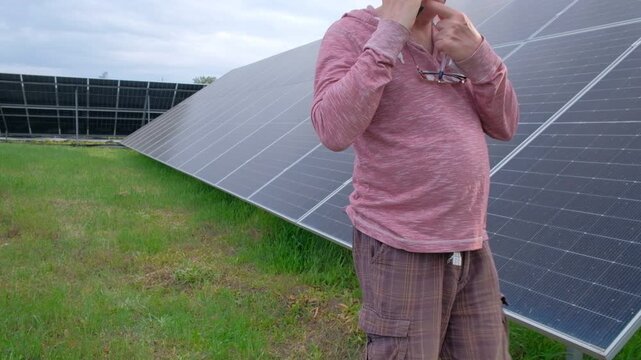 Farmer checking solar panels performance while working in the garden A man with a hoe stands near home solar panels, calculating energy efficiency on a smartphone and taking notes in a notebook. 