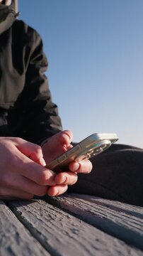Hands type and scroll on a smartphone resting on a wooden bench outdoors against a blue sky. Concept of digital communication, staying connected, and modern mobile technology usage.