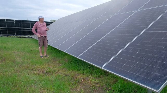 Farmer checking solar panels performance while working in the garden A man with a hoe stands near home solar panels, calculating energy efficiency on a smartphone and taking notes in a notebook. 