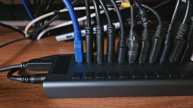 Numerous USB cables are firmly plugged into a long glowing multiport hub resting on a wooden office desk. Concept of digital connectivity, electronic power management, and data transfer.