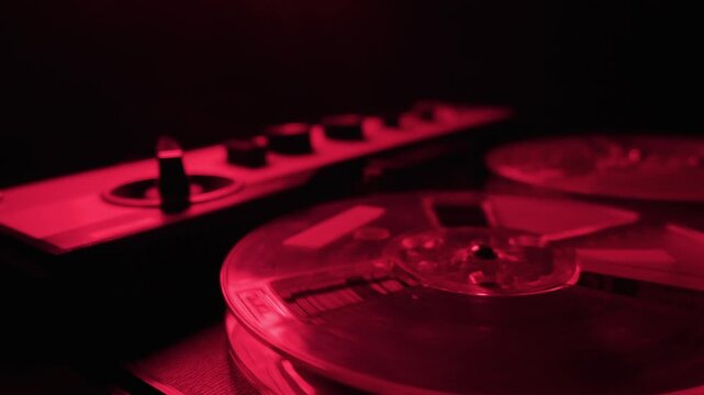 A close-up view of a vintage reel-to-reel tape recorder playing magnetic audio tape. The transparent plastic spools continuously spin while bathed in deep red illumination.