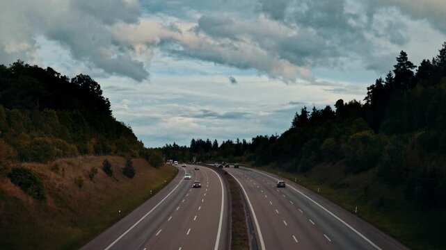 A busy multi-lane highway with moderate traffic including cars and trucks, featuring digital speed limit signs displaying 120, set against a backdrop of dense forest.