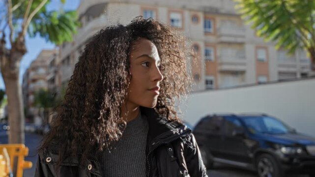 Teenage woman in profile looking right, face and curly hair lit by sunlight, wearing jacket on an urban street; quiet serenity.