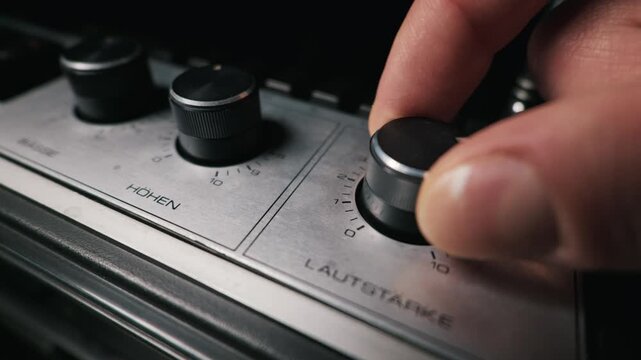 A person hand turns the black volume knob on a vintage silver audio amplifier. The metal front panel features German text for volume and treble. The audio equipment sits in a dark room.