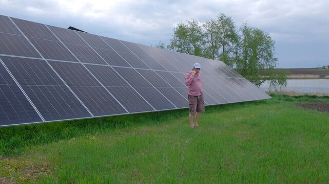 Farmer checking solar panels performance while working in the garden A man with a hoe stands near home solar panels, calculating energy efficiency on a smartphone and taking notes in a notebook. 