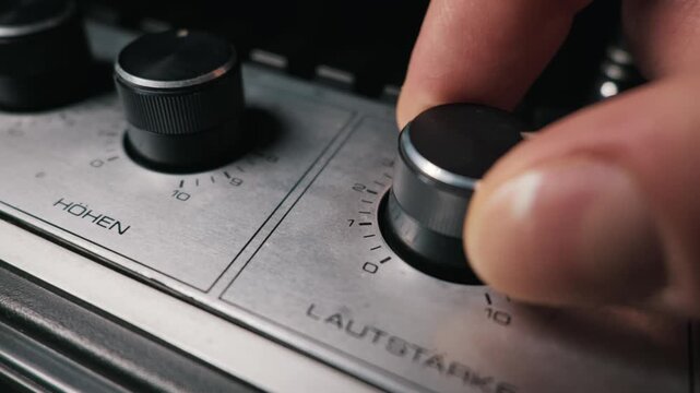 Fingers turn the black volume dial on a brushed metal audio amplifier. The vintage stereo receiver features classic analog controls with German text. A macro view of sound adjustment.