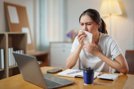 Young Asian woman feeling unwell at home, experiencing cold or flu symptoms, covering her mouth with a tissue while working remotely on a laptop, signifying illness and health issues