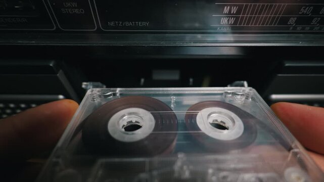 Person inserts a clear audio cassette tape into the open deck of a vintage black stereo boombox. Concept of retro media nostalgia and returning to classic analog music listening experiences.