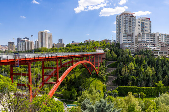 Panoramic view of Dikmen Valley of Ankara featuring a striking red steel arch bridge, lush green terraced parklands, and modern residential high-rises under a sunny blue sky.
