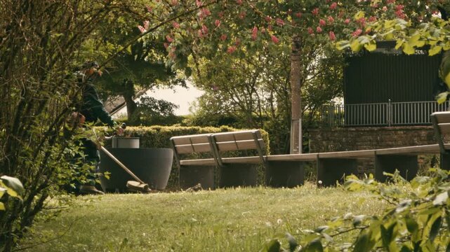 Professional gardener wearing safety gear and a helmet using a string trimmer to cut grass and weeds near park benches and blooming trees on a sunny day.