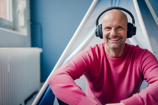 Portrait of smiling mature man wearing headphones sitting at teepee indoors