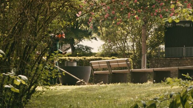 Professional gardener wearing safety gear and a helmet using a string trimmer to cut grass and weeds near park benches and blooming trees on a sunny day.