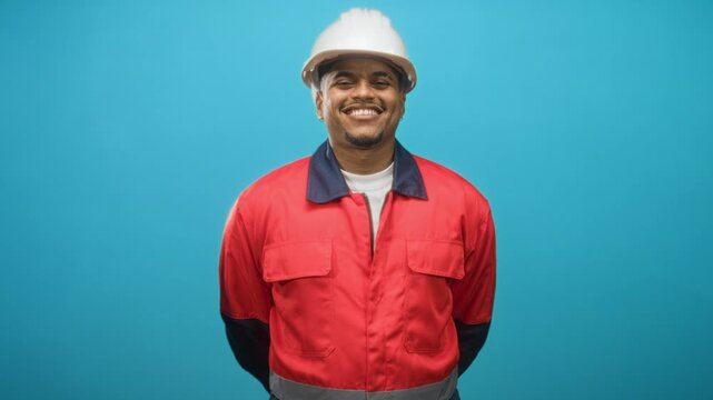 Man in white hard hat and red uniform stands center frame smiling with hands behind back wearing safety coveralls in studio; pride duty.