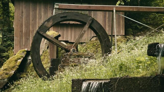A rustic wooden water mill featuring a large, moss-covered water wheel powered by water flowing from a wooden trough, set against a lush green hillside