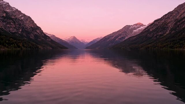 The video features a serene and expansive view of a calm body of water, likely a lake, flanked by towering, rugged mountains on both sides