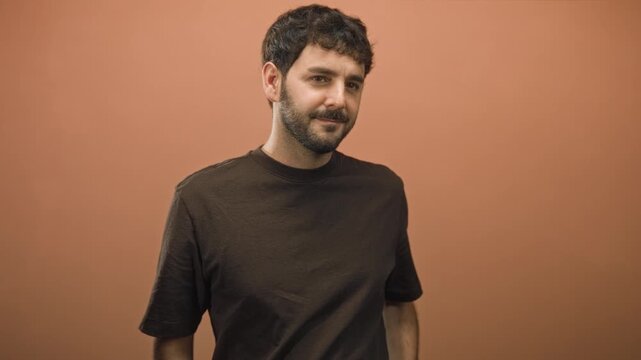 Man with beard crossing arms and slight smile in studio portrait with warm terracotta wall and dark tshirt; quiet confidence.