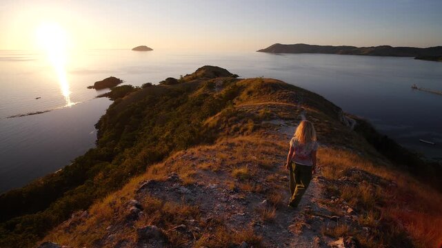 Woman walking along a rugged hilltop trail above Labuan Bajo at sunset, captured in super slow motion with golden light over calm sea, islands, and dramatic tropical coastline