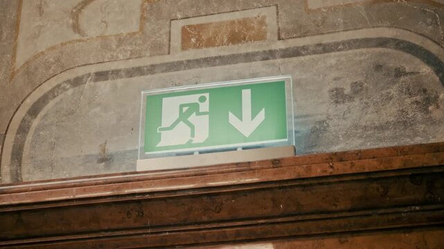 A modern green emergency exit sign with a downward arrow mounted above an antique wooden lintel, set against a backdrop of faded, classical wall frescoes in a historic building.