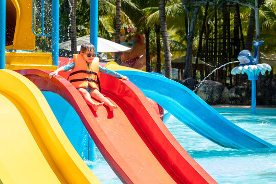 Child boy wearing an orange life jacket joyfully slides down a bright red water slide at tropical water park, with palm trees background. Happy childhood and entertainment concept. High quality photo