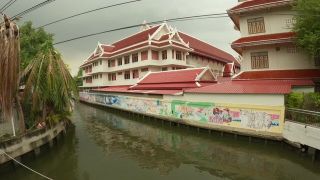 The video footage provides a serene view of Khlong Phasi Charoen in Bangkok, Thailand. The canal is lined with lush green trees, traditional boat structures with covers, and a large, multi-story build