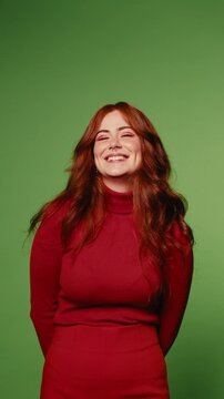A young woman with long, auburn hair smiles and laughs against a vibrant green screen background. She's wearing a stylish red turtleneck, exuding warmth and confidence.