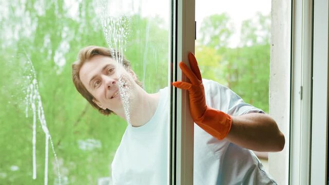Smiling man in orange gloves uses a squeegee to wash a window pane. Happy guy wiping foam during house spring cleaning chores. High quality 4k footage