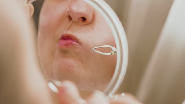 Woman looking in a magnifying mirror, using tweezers for mustache removal. A daily home beauty routine tackling unwanted facial hair problems caused by hormonal changes. High quality 4k footage
