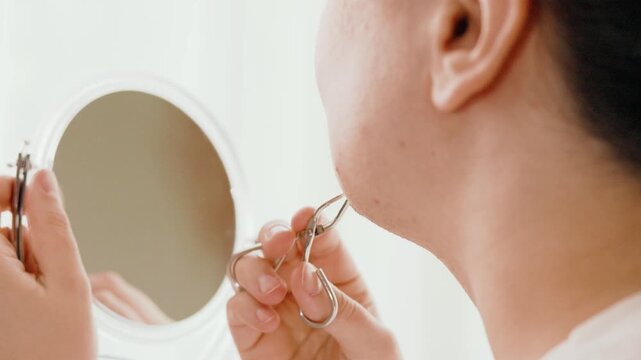 Close-up of a woman removing unwanted facial hair from her chin using tweezers while looking into a mirror. Home beauty routine for hormonal chin hair problems. High quality 4k footage