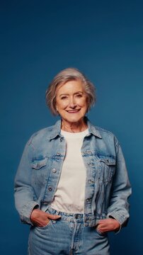 Portrait of a stylish senior woman in denim jacket and jeans on blue background. She exudes confidence and happiness.