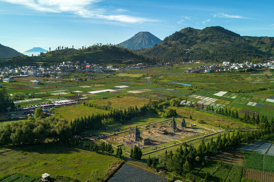 Aerial view of the ancient Dieng temple complex with people walking near the stone structures surrounded by green fields and Mount Sindoro in Kejajar, Central Java, Indonesia.
