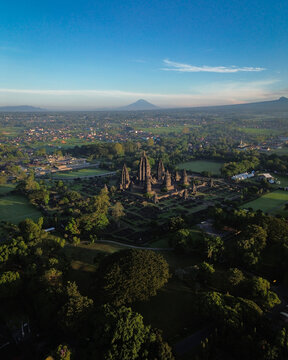 Aerial view of Prambanan Temple complex with stone spires surrounded by lush trees and a distant volcano under blue sky Kecamatan Prambanan, Daerah Istimewa Yogya, Indonesia.