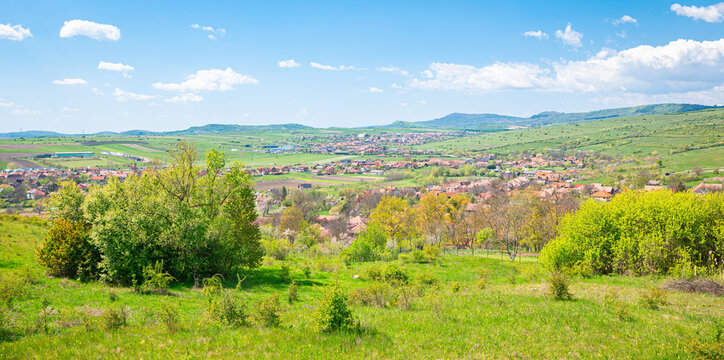 Bright spring landscape of rolling green fields and trees overlooking a small village and distant hills under a clear blue sky, north of Odorheiu Secuiesc, Romania