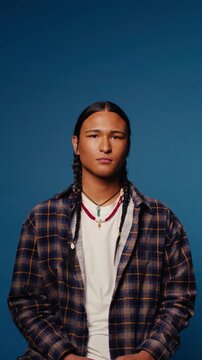Young Indigenous man with braids and a plaid shirt sits calmly against a blue background in a medium close-up vertical shot.