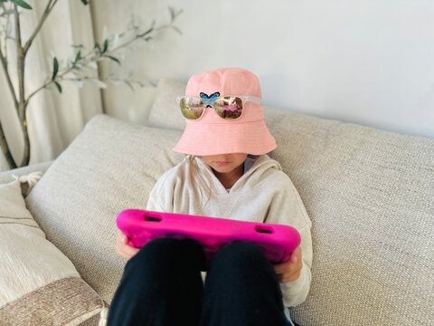 Young Girl wearing a bucket hat and sunglasses sitting on a sofa Playing on a digital Tablet
