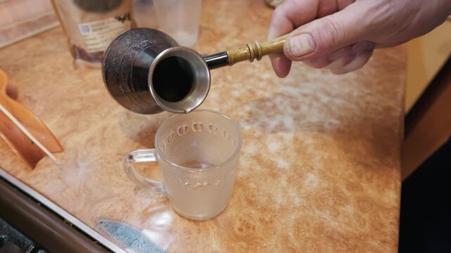 A close-up view shows a hand holding a traditional copper cezve coffee pot above a clear glass mug on a kitchen counter. In the background, a bag of roasted beans sits next to kitchenware.