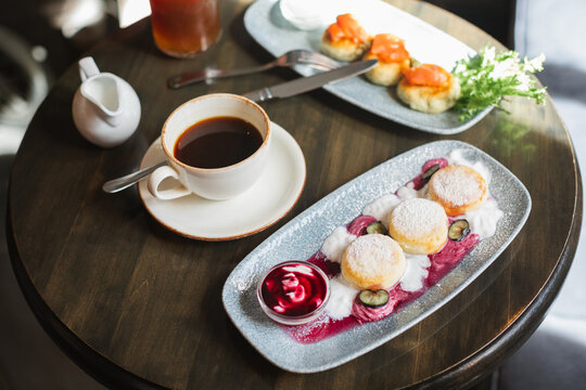 Overhead view of a plate of cottage cheese pancakes (syrniki) with cream and blueberry jam, a cup of coffee and cottage cheese pancakes with smoked salmon on a table