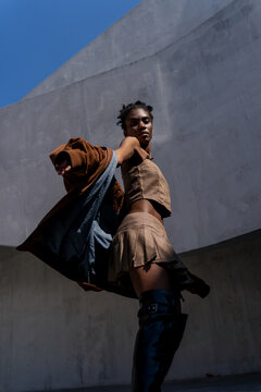 Low angle side view of a stylish Black woman in a brown mini skirt and leather boots messing about in front of a concrete brutalist wall