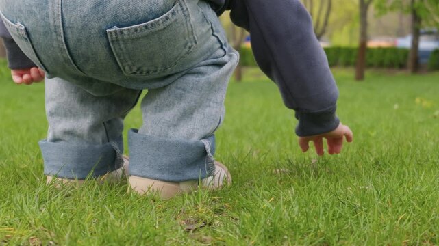 A young child dressed in denim overalls is captured squatting on a vibrant green lawn. The child is actively reaching out to explore the grassy surface in a sunlit public park. This scene