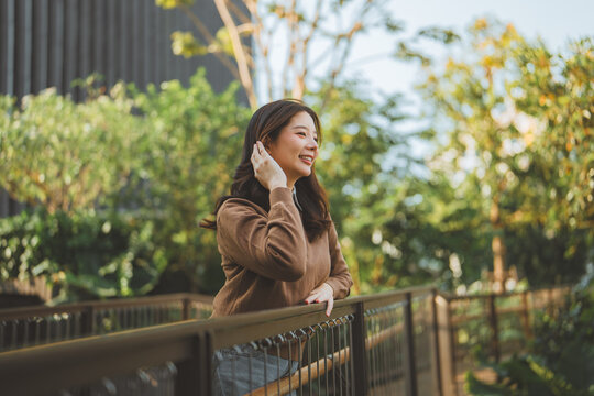 Happy young Asian woman standing on balcony bridge and smiling while looking away, Cheerful female relaxing in green garden park, outdoor leisure activity, healthy lifestyle and positive thinking