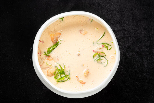 A white ceramic bowl filled with creamy coconut soup featuring shrimp, fresh green herbs, and scallions, set against a black background.