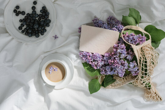 Overhead view of a blank deckled edge card in a string bag with fresh lilac flowers, a cup of coffee and plate of blueberries on crumpled bed linen