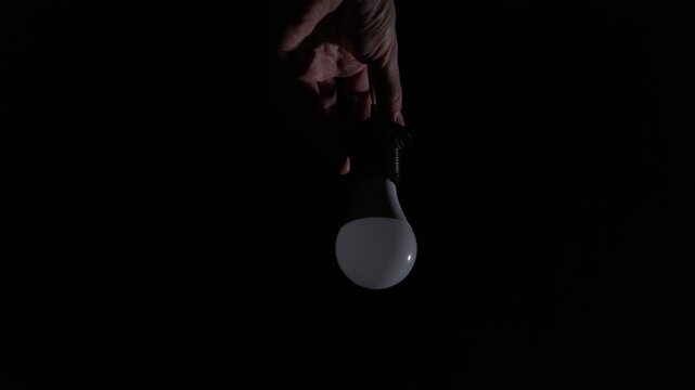 Hands carefully unscrew a glowing LED light bulb from a hanging black socket. The bright bulb flickers slightly against the completely dark background. A hand reaches up from below.