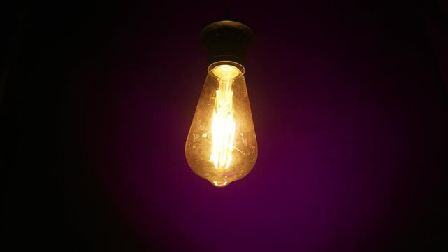 A glowing filament light bulb hangs suspended from a dark ceiling fixture. The illuminated glass bulb casts a warm yellow light against a deep purple background in a shadowed interior space.