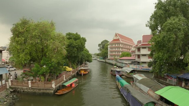 The video footage provides a serene view of Khlong Phasi Charoen in Bangkok, Thailand. The canal is lined with lush green trees, traditional boat structures with covers, and a large, multi-story build