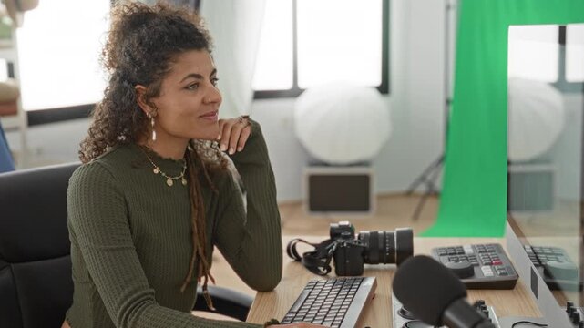 Young woman resting chin on hand at keyboard beside camera and green screen in studio; thoughtful creativity.
