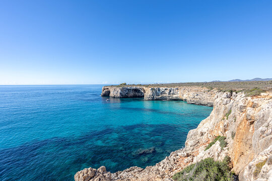 Crystal clear turquoise Mediterranean water along rugged limestone cliffs near Cala Varques, with a natural rock arch and small boat anchored in the secluded bay