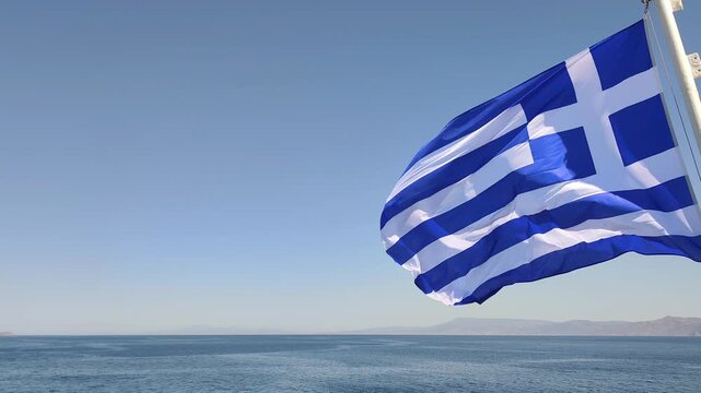 View of the Greek national flag billowing in the sea breeze on a ferry crossing the Aegean Sea, set against a clear summer-blue sky.