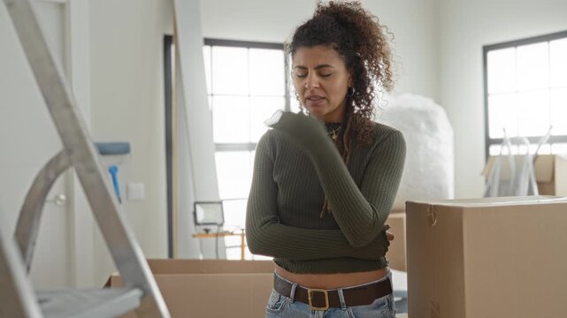 Woman rubbing her eye with hand, arms folded, standing amid cardboard moving boxes, ladder and ironing board in a bright building interior; moving stress.
