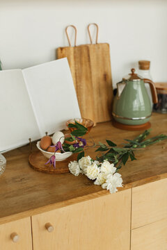 Close-up of an open book with blank pages, a kettle, wooden chopping boards, fresh eggs and flowers on a  wooden kitchen worktop